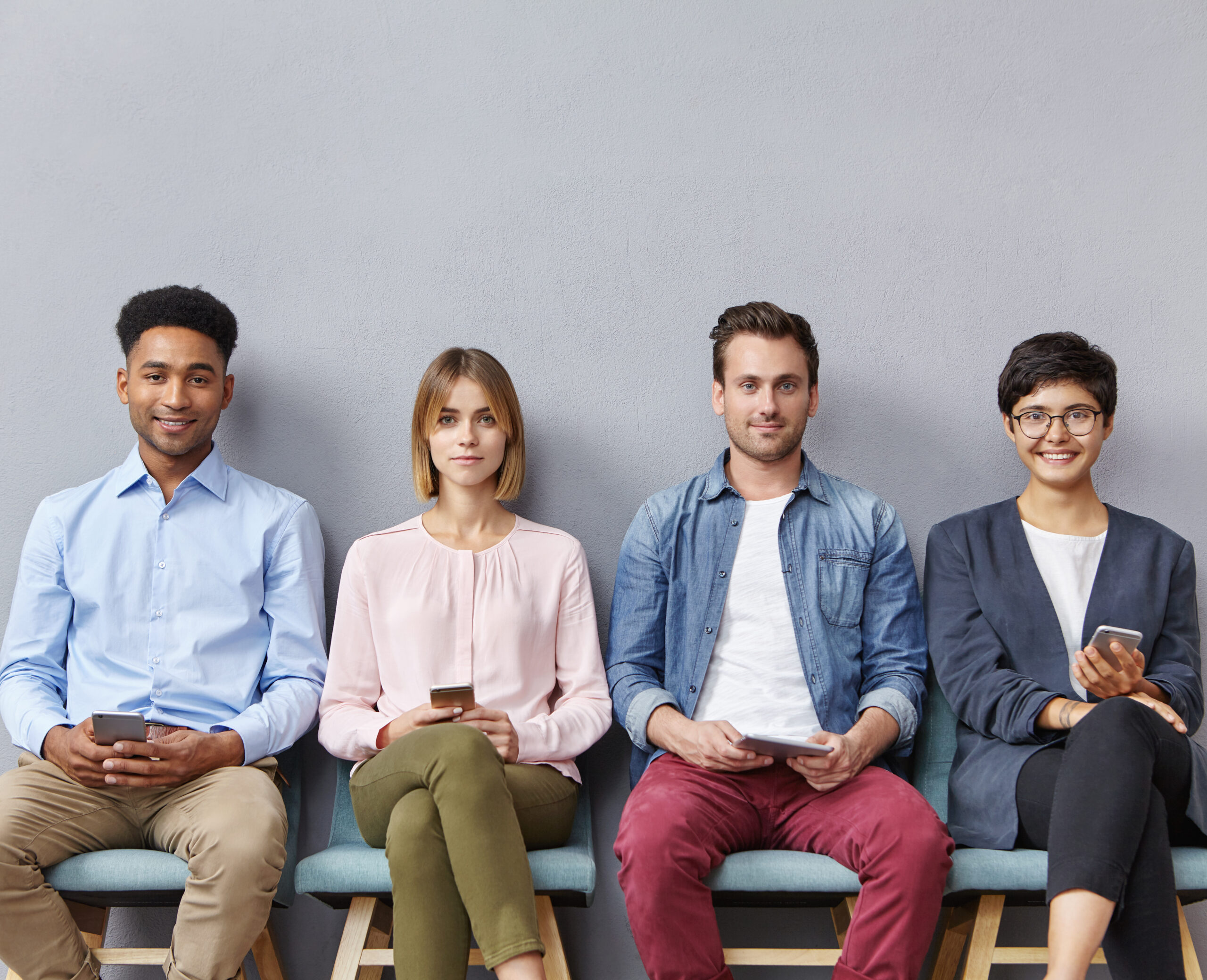 Technology, ethnicity and people concept. International group of young women and men sit in queue line with modern smartphones and tablets, wait for job interview, isolated over grey blank wall.